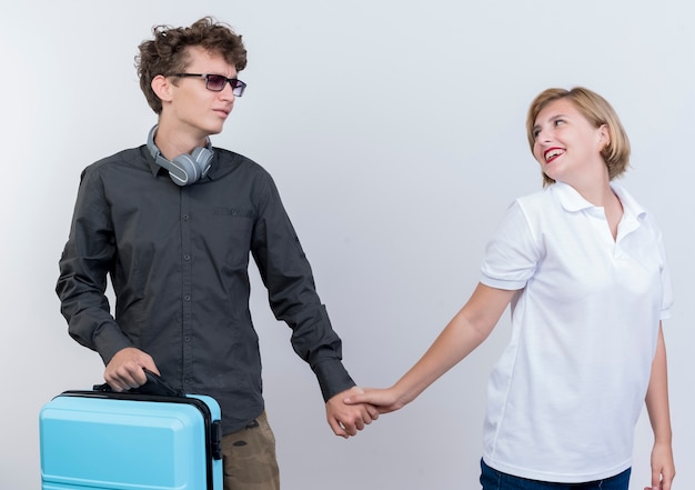 Jeune couple homme avec valise laissant sa petite amie heureuse toucher sa main debout sur un mur blanc