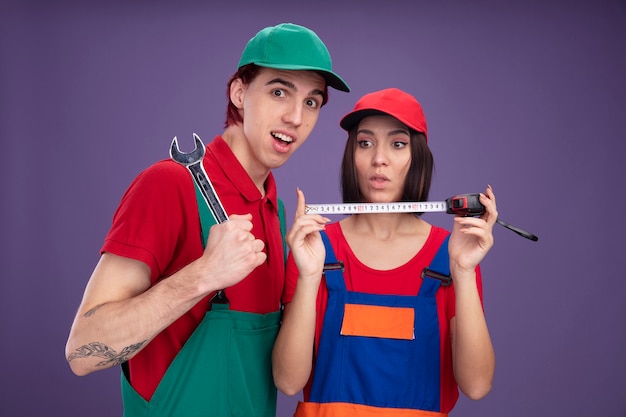 Jeune couple impressionné en uniforme de travailleur de la construction et fille de casquette tenant et regardant un gars de mètre à ruban tenant une clé regardant la caméra isolée sur un mur violet