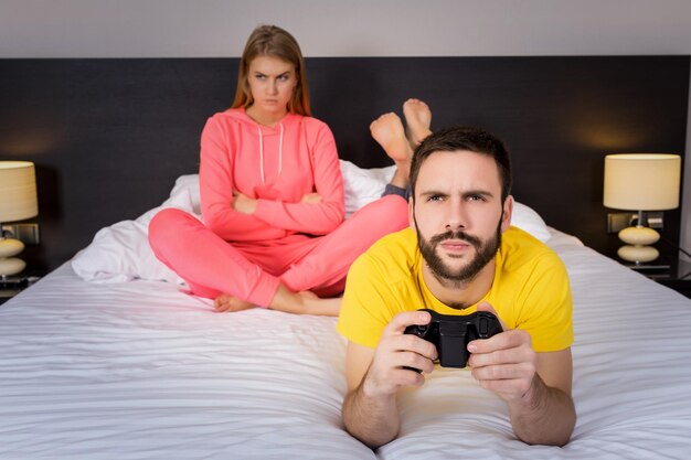 Jeune couple jouant à des jeux vidéo au lit. Homme jouant au jeu vidéo, femme bouleversée en arrière-plan.