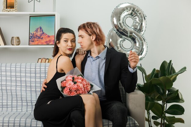 Jeune couple le jour de la femme heureuse avec bouquet assis sur un canapé dans le salon