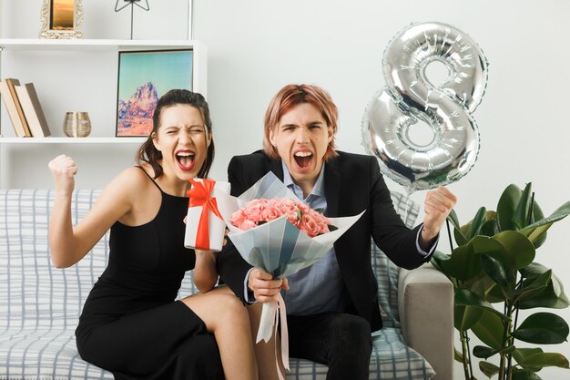 Jeune couple le jour de la femme heureuse tenant présent avec bouquet assis sur un canapé dans le salon