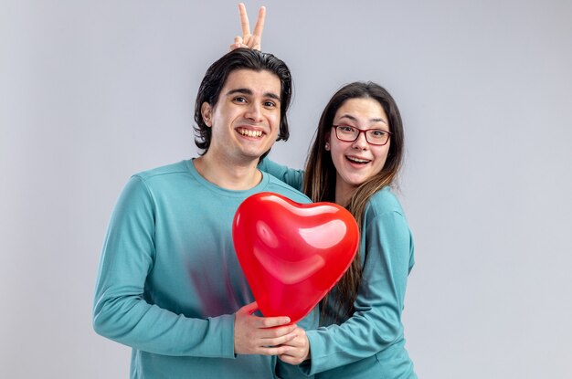 Jeune couple le jour de la Saint-Valentin fille souriante faisant un geste d'oreilles de lapin pour gars avec ballon coeur isolé sur fond blanc