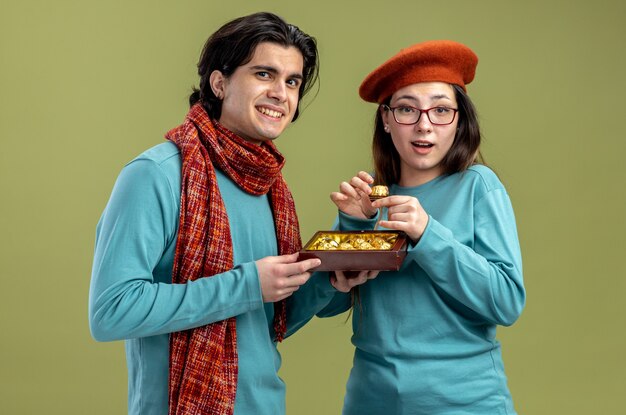 Jeune couple le jour de la Saint-Valentin gars portant une écharpe fille portant un chapeau mec souriant donnant une boîte de bonbons à une fille heureuse isolée sur fond vert olive