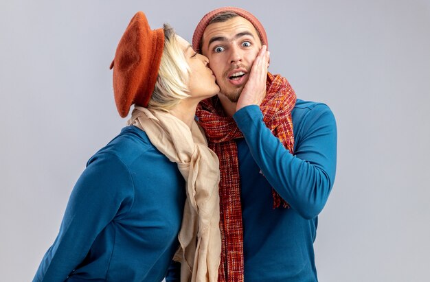 Jeune couple le jour de la Saint-Valentin portant un chapeau avec une écharpe fille heureuse embrassant un gars surpris isolé sur fond blanc