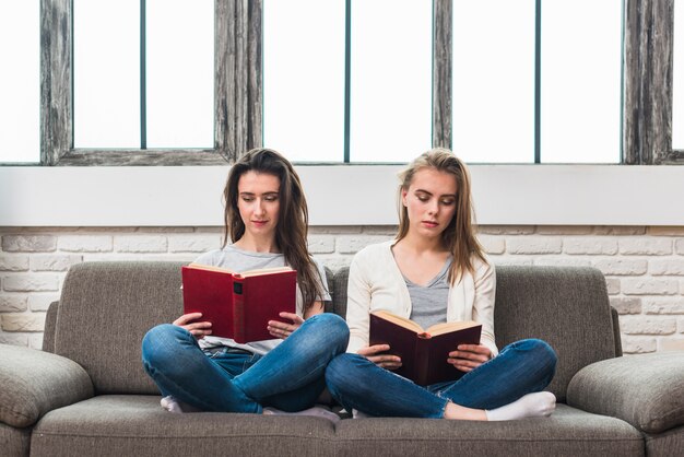 Jeune couple de lesbiennes assis sur un canapé gris avec livre de lecture jambes croisées