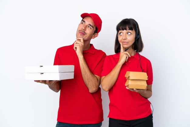 Jeune couple de livraison tenant des pizzas et des hamburgers isolés sur fond blanc en pensant à une idée tout en levant les yeux