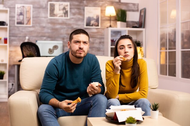 Jeune couple mangeant du poulet frit devant la télévision dans le salon