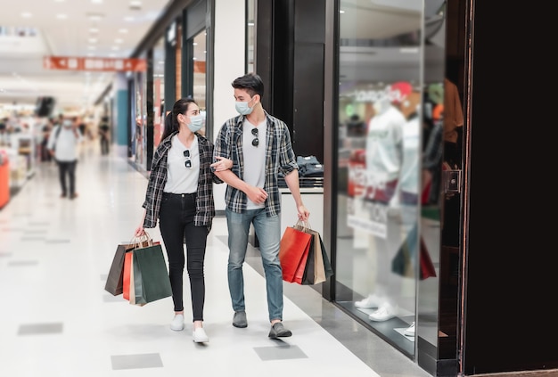 Jeune couple en masque de protection tenant plusieurs sacs en papier marchant dans le couloir du grand centre commercial