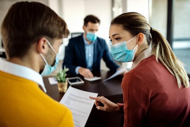 Jeune couple avec des masques faciaux discutant de la paperasse lors d'une réunion avec leur conseiller financier