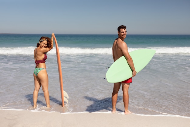 Jeune couple avec planche de surf en regardant la caméra sur la plage au soleil