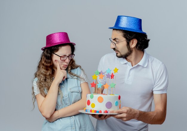 Jeune couple portant des chapeaux roses et bleus guy donne un gâteau d'anniversaire à une fille confuse isolée sur blanc