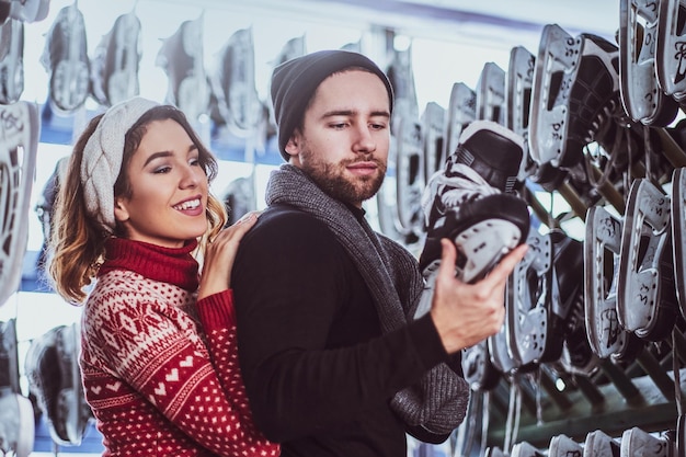 Jeune couple portant des vêtements chauds debout près du rack avec de nombreuses paires de patins, choisissant sa taille