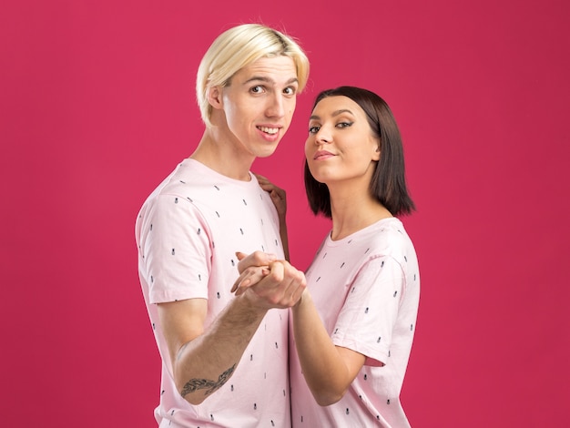 Jeune couple en pyjama debout dans la vue de profil homme impressionné et femme confiante tenant la main regardant à l'avant isolé sur un mur rose avec espace de copie