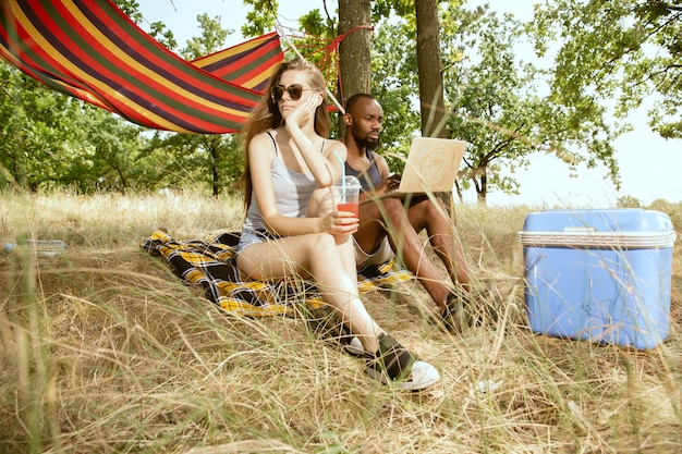 Jeune couple romantique international multiethnique à l'extérieur à la prairie en journée d'été ensoleillée. Homme afro-américain et femme caucasienne ayant pique-nique ensemble. Concept de relation, l'été.