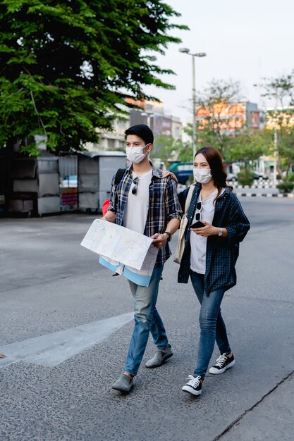 Jeune couple de routards asiatiques en masque de protection contre le coronavirus et vérifier l'emplacement sur une carte papier tout en marchant dans la rue de la ville, Jolie femme tenant un smartphone à la main