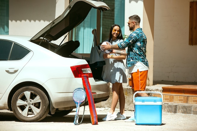 Jeune couple se préparant pour un voyage de vacances sur la voiture en journée ensoleillée. Femme et homme empilant des équipements de sport. Prêt pour la mer, la rivière ou l'océan. Concept de relation, été, week-end.