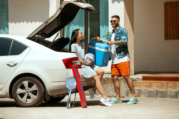 Jeune couple se préparant pour un voyage de vacances sur la voiture en journée ensoleillée. Femme et homme empilant des équipements de sport. Prêt pour la mer, la rivière ou l'océan. Concept de relation, été, week-end.