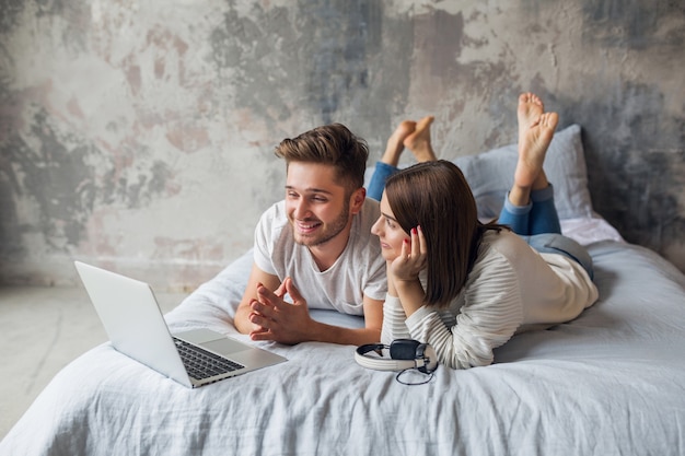 Jeune couple souriant allongé sur le lit à la maison en tenue décontractée, à la recherche d'un ordinateur portable, homme et femme, passer du bon temps ensemble, se détendre