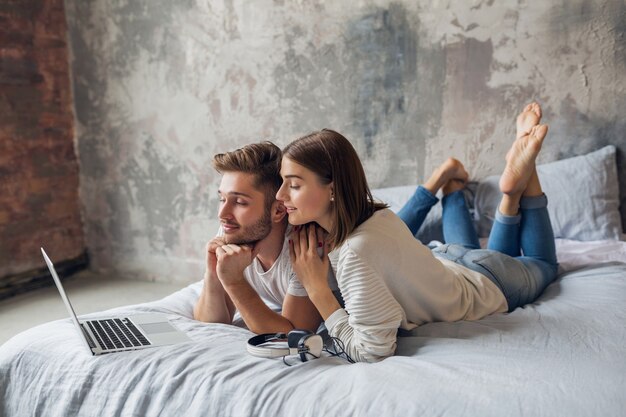 Jeune couple souriant allongé sur le lit à la maison en tenue décontractée, à la recherche d'un ordinateur portable, homme et femme, passer du bon temps ensemble, se détendre