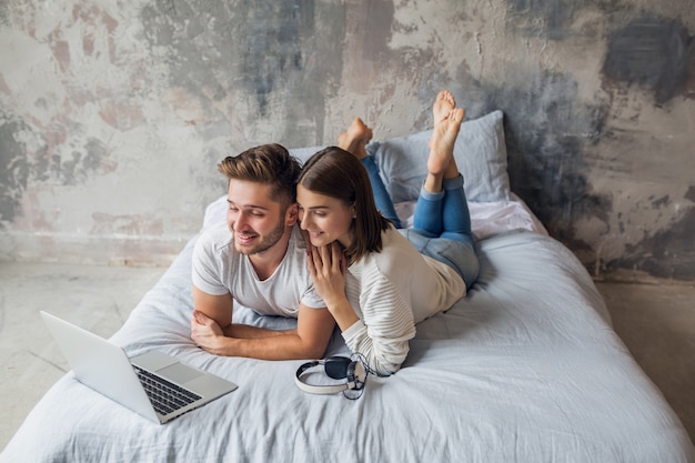 Jeune couple souriant allongé sur le lit à la maison en tenue décontractée, à la recherche d'un ordinateur portable, homme et femme, passer du bon temps ensemble, se détendre