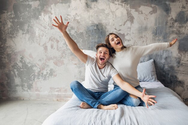 Jeune couple souriant assis sur le lit à la maison en tenue décontractée, homme et femme s'amusant ensemble, émotion positive folle, heureux, tenant les mains