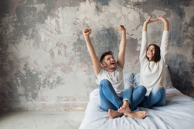 Jeune couple souriant assis sur le lit à la maison en tenue décontractée, homme et femme s'amusant ensemble, émotion positive folle, heureux, tenant les mains