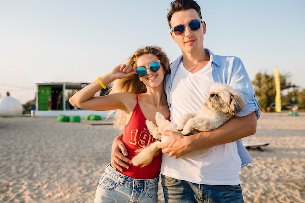 Jeune couple souriant attrayant s'amusant sur la plage en jouant avec le chien de race shih-tsu