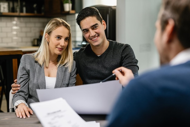 Jeune couple souriant ayant une réunion avec un agent immobilier à la maison et parlant de nouveaux plans de logement