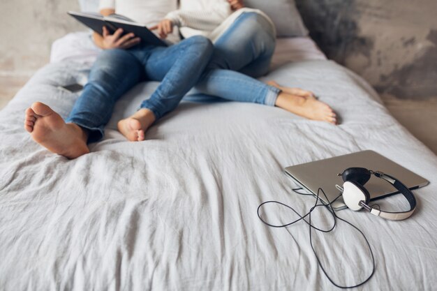 Jeune couple souriant heureux assis sur le lit à la maison en tenue décontractée livre de lecture portant des jeans, homme et femme jambes humeur romantique, ordinateur portable