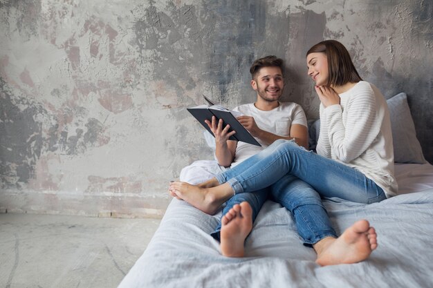 Jeune couple souriant heureux assis sur le lit à la maison en tenue décontractée, livre de lecture portant des jeans, homme et femme, passer du temps romantique ensemble