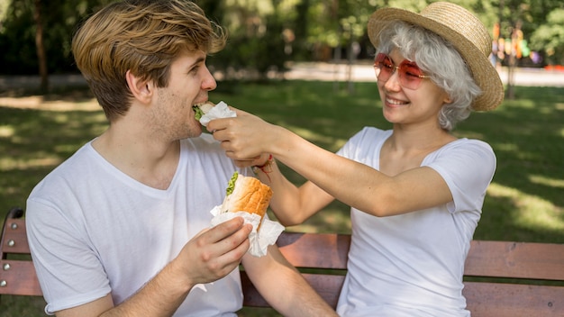 Jeune couple souriant, manger des hamburgers dans le parc