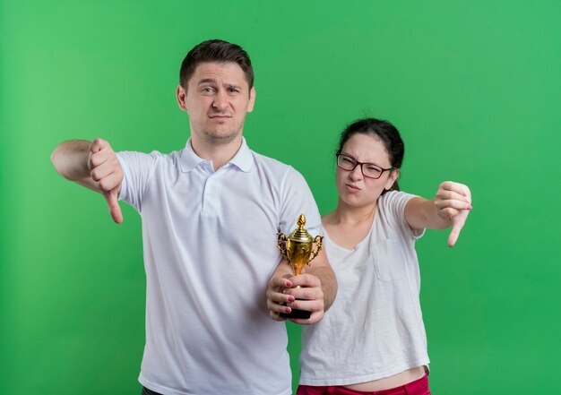 Jeune couple sportif homme et femme debout ensemble tenant le trophée montrant les pouces vers le bas debout sur le mur vert