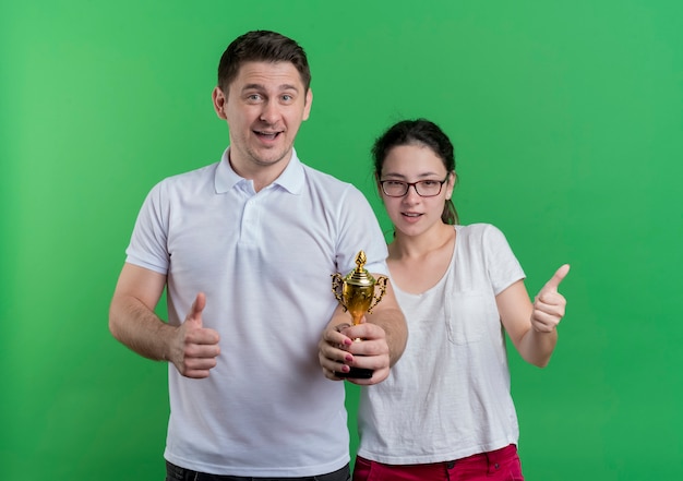 Jeune couple sportif homme et femme debout ensemble tenant le trophée montrant les pouces vers le haut debout sur le mur vert