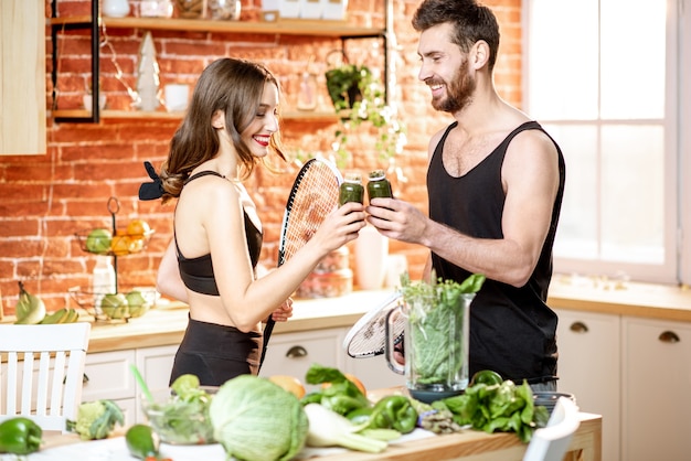 Jeune couple sportif prenant une collation avec de la nourriture végétarienne buvant un smoothie debout avec des raquettes dans la cuisine à la maison