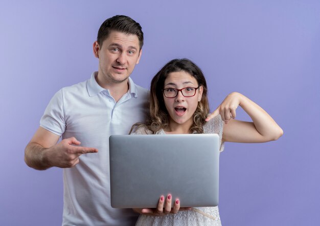 Jeune couple surpris femme ordinateur portable hlding pointant avec le doigt à côté de son petit ami sur bleu