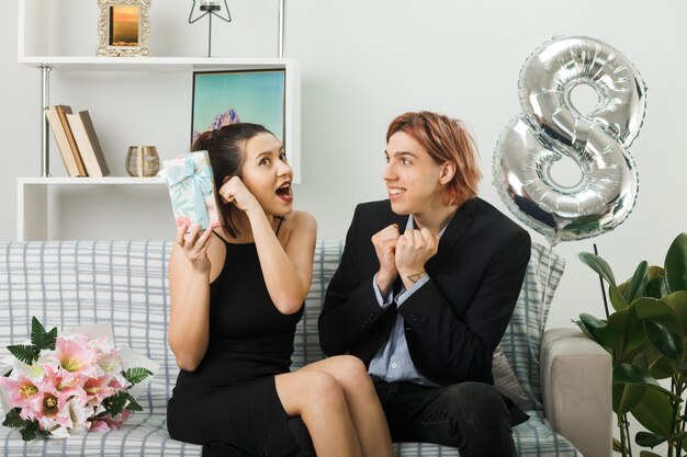 Jeune couple surpris lors de la journée des femmes heureuses tenant un mec présent montrant un geste oui assis sur un canapé dans le salon