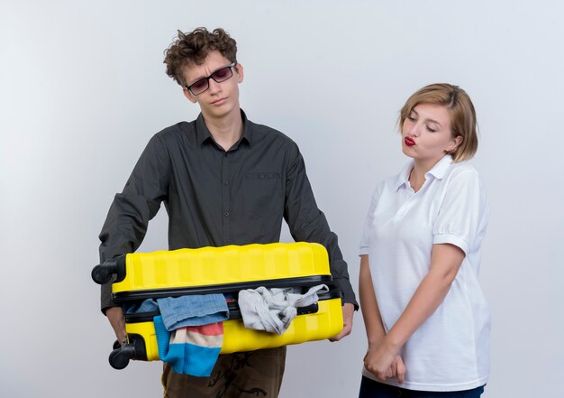 Jeune couple de touristes homme tenant une lourde valise pleine de vêtements fatigués debout à côté de sa petite amie avec une expression triste sur un mur blanc