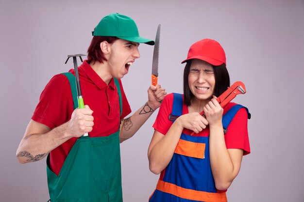 Jeune couple en uniforme de travailleur de la construction et casquette fille effrayée tenant une clé à pipe avec les yeux fermés mec en colère tenant un râteau et une scie à main regardant une fille criant