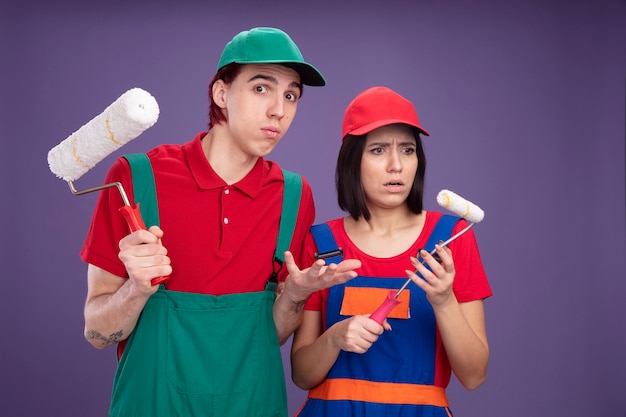 Jeune couple en uniforme de travailleur de la construction et casquette tenant un rouleau à peinture mec ignorant regardant la caméra montrant une main vide fille confuse regardant son rouleau à peinture isolé sur un mur violet