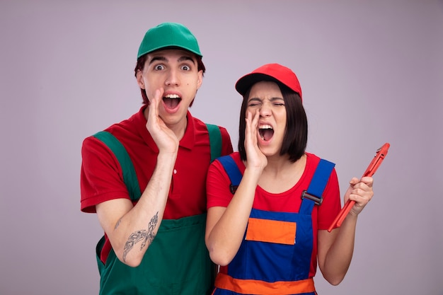 Jeune couple en uniforme de travailleur de la construction et chapeau fille furieuse tenant une clé à pipe criant à haute voix avec les yeux fermés mec excité les deux gardant la main près de la bouche