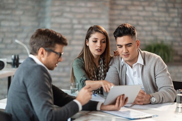 Jeune couple utilisant un pavé tactile avec un agent d'assurance et tout en faisant un plan de remboursement de prêt lors d'une réunion