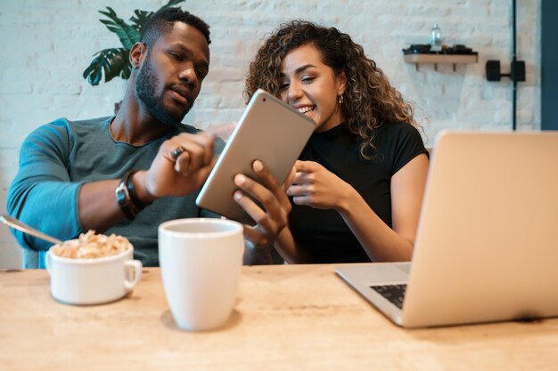 Jeune couple utilisant une tablette numérique et un ordinateur portable tout en prenant le petit déjeuner ensemble à la maison.