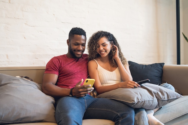 Jeune couple utilisant un téléphone portable assis sur un canapé à la maison.