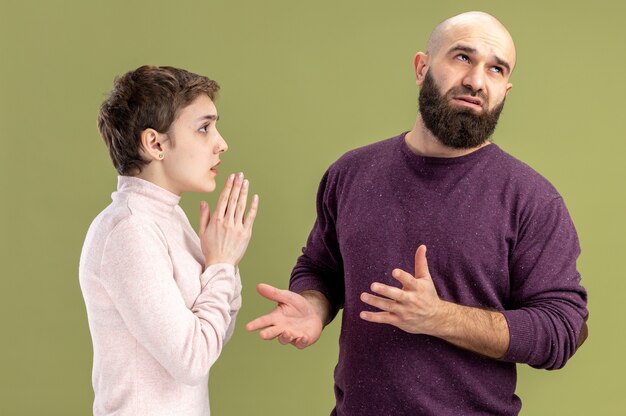 Jeune couple en vêtements décontractés femme aux cheveux courts demandant pardon son petit ami barbu confus Saint Valentin concept debout sur mur vert