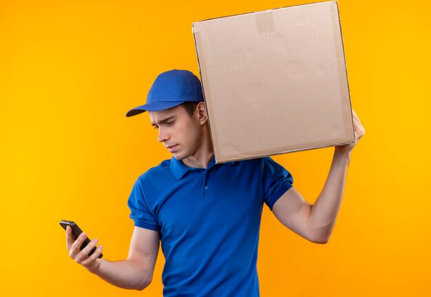 Jeune courrier portant l'uniforme bleu et bonnet bleu tenant une boîte tient un téléphone