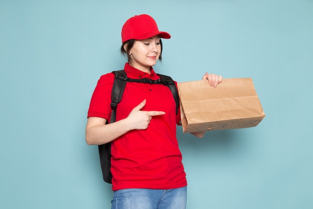 Jeune coursier en polo rouge casquette rouge sac à dos noir tenant le paquet souriant sur bleu