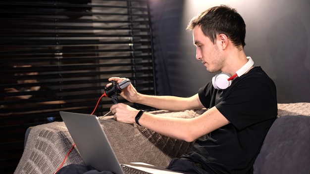 Un jeune créateur de contenu installe le microphone, l'ordinateur portable à genoux, assis sur le canapé. Travailler à domicile