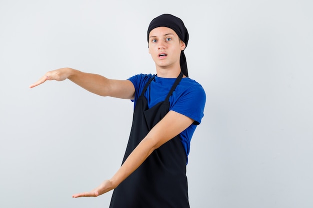 Jeune cuisinier adolescent montrant un signe de grande taille en t-shirt, tablier et l'air perplexe, vue de face.