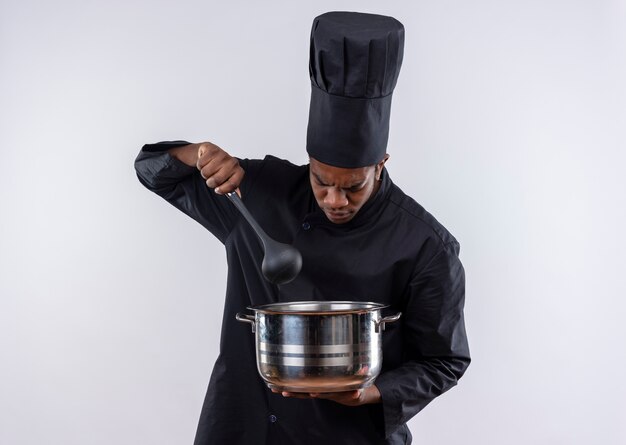 Jeune cuisinier afro-américain agacé en uniforme de chef détient casserole et cuillère isolé sur fond blanc avec espace copie
