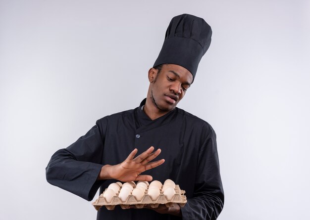 Jeune cuisinier afro-américain agacé en uniforme de chef détient lot d'oeufs et fait semblant de repousser isolé sur fond blanc avec copie espace